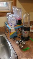 Magic Bullet blender base with tall clear blending cup attached, several additional blending cups and lids, and small metal accessories on countertop near sink.