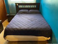 Full bedframe with mattress and dark quilted cover in a room with blue walls and carpet floor.
