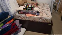 Photo showing side view of dark platform bedframe supporting a floral-patterned mattress with folded linens and cushions on top.