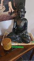 Black Buddha statue seated on a woven mat with a brown pillar candle on a wooden surface.