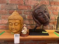 Three items in the lot displayed on a shelf against a brick wall: glazed ceramic Buddha bust, dark Buddha head sculpture on stand, and small figurine on wood base