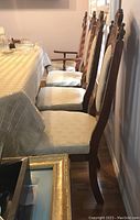 Side view of four dining chairs at a table, three without arms and one with wooden arms, upholstered seats and backs in cream patterned fabric.