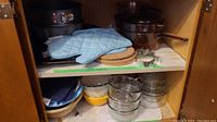 Cabinet shelf showing stacked glass saucepans with handles, springform pans, oven mitts, wood trivets, and various bowls beneath.