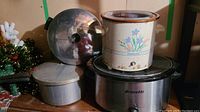 Electric stainless steel crock pot with black base and glass lid, brand Bravetti, shown with vintage enamel crock pot on top.