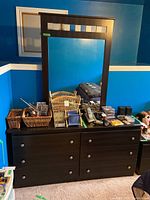 Front view of black laminate dresser with six drawers and attached mirror. Items placed on top include wicker baskets and personal belongings.