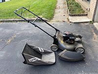 Side view of Craftsman gas lawn mower with grass catcher bag and side discharge chute placed beside it on asphalt driveway.