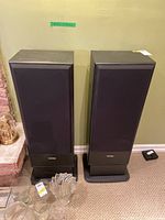 Pair of black floor-standing speakers with black grille covers, side wood panel finish, and wide base stands placed on carpet against a green wall.