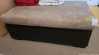 Front and top view showing beige cushioned top and dark brown base of storage bench with some bedding on top.
