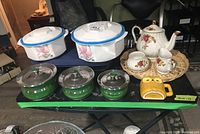 Photo showing white casserole dishes with blue-rimmed lids, green pots with lids, porcelain teapot set and yellow owl cup on wicker tray