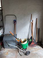 Photo shows metal seed spreader cart with attached green plastic bucket, assorted yard tools including large metal snow shovels, leaf rakes with wooden and plastic handles, and smaller handheld tools.