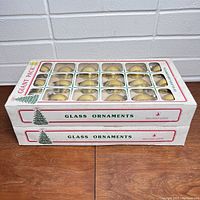 Two unopened white boxes each containing 18 gold glass Christmas ornament balls visible through plastic windows. Boxes stacked and shown from the front and side.