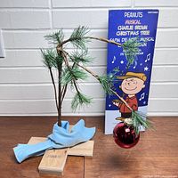 Charlie Brown sparse pine branch Christmas tree with single red ornament and blue fabric wrap base, wooden cross stand, against brick background.