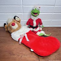 Kermit the Frog plush in Santa outfit standing beside Rudolph plush stocking laying on floor, showing both items together.