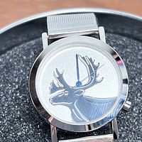 Close-up of men's wristwatch dial showing Inuit style moose head graphic in black on white dial with silver bezel and metal mesh band.