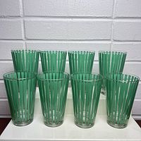Front view of 8 vintage glasses arranged in two rows on a white surface with a white brick background. Glasses are clear with vertical green stripes and silver accents.