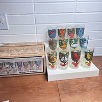 12 colorful 12 Days of Christmas Brockway glasses arranged in a display stand next to the original box showing artwork of all 12 glass designs.