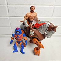 Two vintage Masters of the Universe figures, a muscular male on a mechanical horse with silver armor and weapon, and a smaller blue-orange armored figure with a helmet.