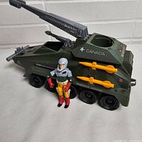 Green GI Joe Persuader armored vehicle with mounted grey cannon and orange missiles, Backstop figure standing beside it.