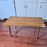 Front angle view of vintage Nasco slatted wood bench table showing rectangular top with six wooden slats and four tapered legs standing on reddish-brown hardwood floor near white kitchen cabinets.