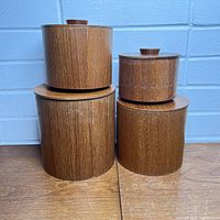 Four vintage teak laminate stackable kitchen canisters with solid wood lids shown assembled and stacked on a wooden surface in front of a blue brick wall.