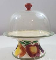 Front view of the ceramic pedestal cheese plate with glass dome showing painted fruit designs (apple, pear) on the base and dome top with wooden knob.