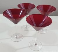 Four martini glasses with deep red bowls and clear stems displayed on a white surface, showing side angles and reflections.