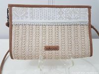 Front view of sakroots handbag showing woven cream fabric with taupe leather edges and decorative band with a zipper top closure.