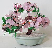 Front view showing pink glass blossoms, green leaves, brown trunk in light green ceramic pot with white gravel.