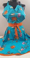 Full front view of turquoise floral apron displayed on dress form, showing overall design, colors, and orange waist tie