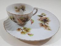 Porcelain teacup on matching snack plate, showing pine needle design and scalloped edges with gold trim