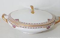 Oval white porcelain serving bowl with lid decorated with pink floral pattern and gold accents on handles and lid knob, shown from angled top.