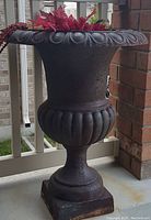 Full view of the large cast iron planter on a porch, showing the overall shape and decorative top edge with faux floral arrangement inside.