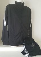 Front view of black men's zip-up jacket with three white stripes on arms, displayed on mannequin with matching pants folded beside it.