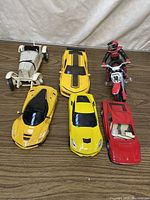 Five collectible model vehicles displayed on a wooden surface: one vintage white race car, three yellow sports cars, one red convertible, and one red motorcycle with rider figurine.
