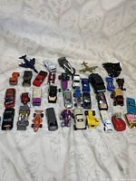 Image showing multiple small die cast toy vehicles including cars, trucks, airplanes, arranged on a white cloth background.