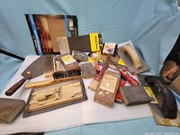 Photo showing a large variety of used hand tools including sharpening stones, wooden tools, brushes, hand saw handle, putty knives, and sanding blocks.