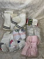 Photo showing multiple packaged socks including white ribbed crew socks with black stripes, baby socks with blue, green, red accents, and pink ribbed socks