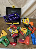 Photo of assortment of colorful plastic toy play tools including drills, hammers, wrenches, and a yellow and black plastic tool box with purple tray.