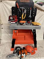 Front view of Black and Decker toy work bench with mounted pegboard and assorted toy tools arranged on bench surface and hanging from hooks, including saw, screwdriver, pliers, wrench, and small accessories.