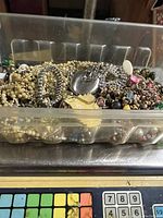 Clear plastic container filled with mixed jewelry items including chains, beads, and pendants.