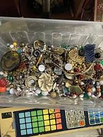 Clear plastic bin containing mixed vintage to modern broken and unmatched jewelry, beads, and findings, showing varied colors, metals, and materials.