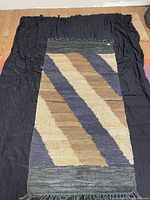Full view of the handwoven leather chindi rug on black fabric background showing diagonal stripes in beige, brown, and blue colors with grey fringed edges.