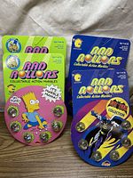 Two unopened Rad Rollers packs: one Simpsons-themed with Bart Simpson graphic and green top border, one Batman-themed in blue packaging.