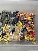 Plastic storage bin containing a mix of vintage and modern toy soldiers, small plastic animals, and military toy vehicles including tanks and jets.
