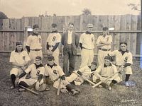 Close-up view of the original 1905 black and white baseball team photograph showing eleven players and one person in a suit, sitting and standing on the grass with bats and gloves, in front of a wooden fence.