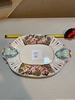 Top view of large oval porcelain platter showing semi-nude cherub women hand-painted design on two sides, gold trim, and blue decorative handles; measuring tape for scale.