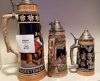 Three vintage West German lidded beer steins of varying sizes displayed together. The largest stein has a man in traditional dress and GERSTENSAFT text, the middle has a town scene, and the smallest has a figurative design.