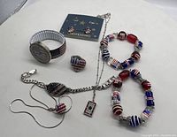 Photo of all items in the lot arranged on a white surface including bracelets, ring, earrings, necklaces, and watch with patriotic red, white, and blue theme