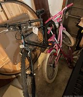 Two bicycles and a pink scooter leaned against a wall in a garage, adult Schwinn mountain bike black in color, youth pink bicycle, and pink Razor scooter.