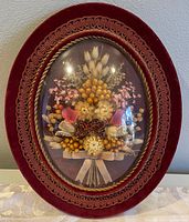 Front view of the oval dried flower artwork featuring yellow, white, and pink dried flowers in a red velvet frame with gold trim.
